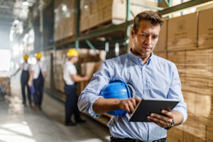 Mid adult foreman surfing the Internet on touchpad in a distribution warehouse. There are people in the background.